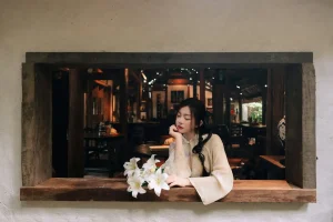 Áo dài sáng màu và bó hoa trắng bên ô cửa gỗ