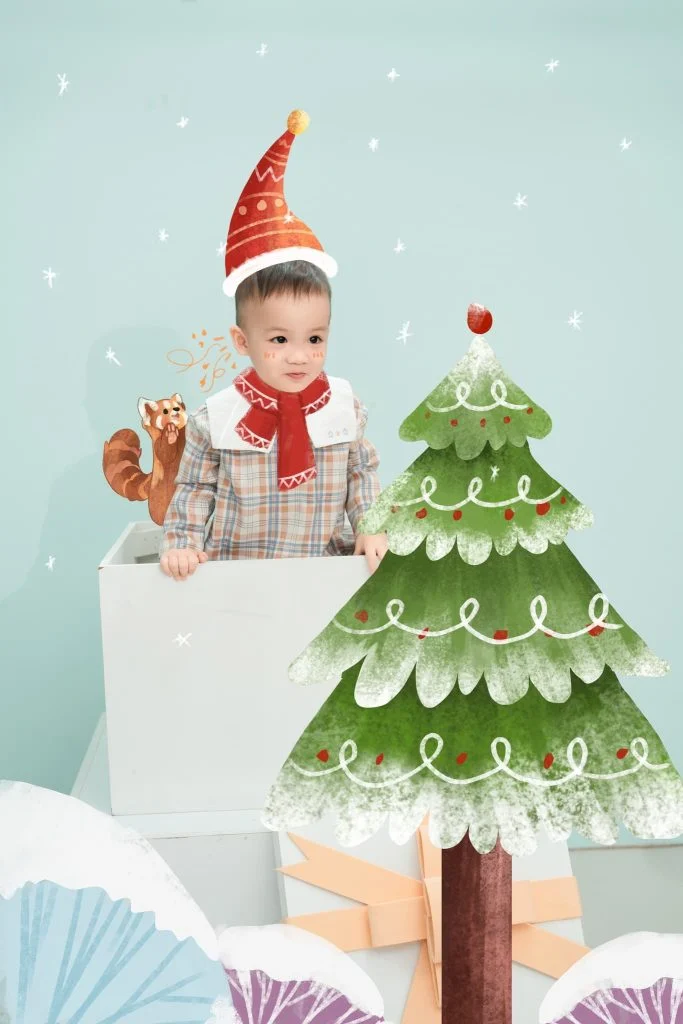 Bé đội mũ sinh nhật ngồi bên cây thông trang trí