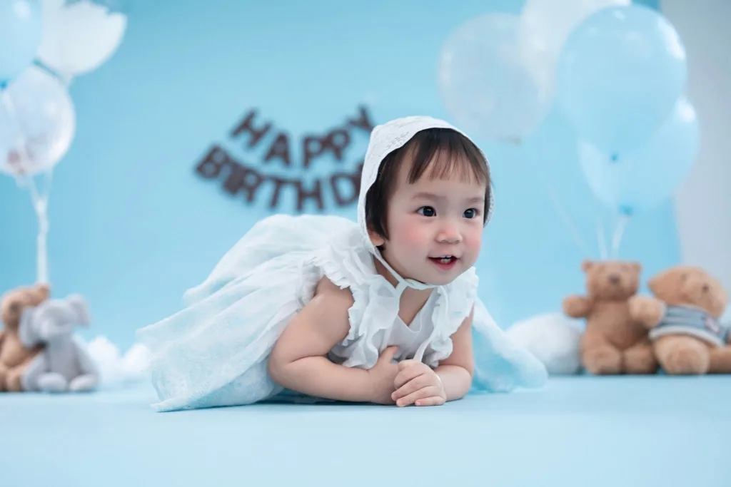 Bé mặc váy trắng nằm chống tay trong bối cảnh sinh nhật xanh