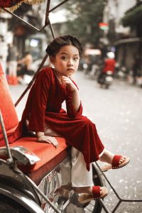 Bé mặc áo dài đỏ ngồi trong bối cảnh ngoài trời