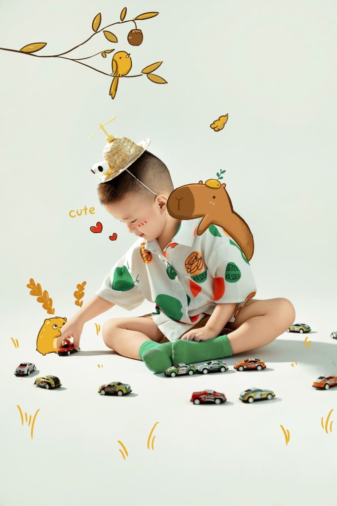 Bé ngồi chơi với những chiếc xe nhỏ trên nền sáng có hình vẽ cây và thú