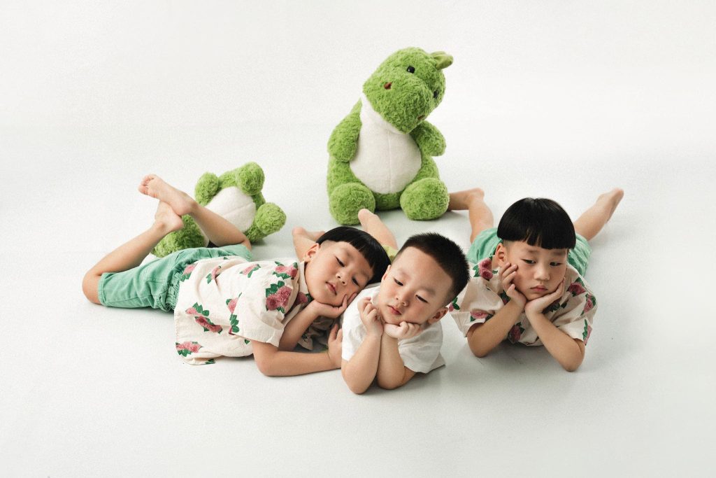 Ba bé nằm sát nhau cùng ôm thú bông trên sàn trắng