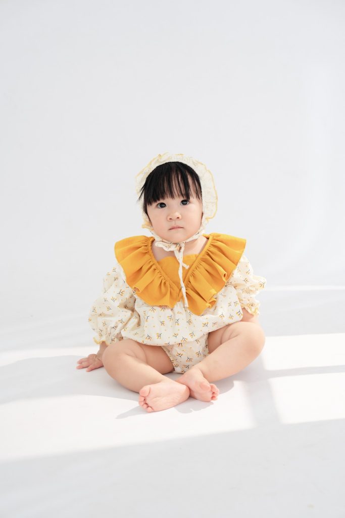 Bé đội mũ trắng, mặc đồ hoa ngồi trên nền sáng