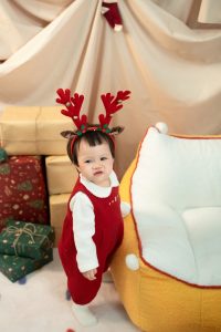 Bé mặc bộ đồ đỏ trắng và bờm tuần lộc bên ghế cùng hộp quà