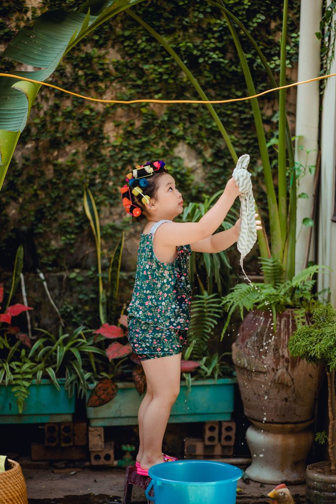 Bé đứng nâng tấm khăn ướt trong bộ đồ hoa xanh