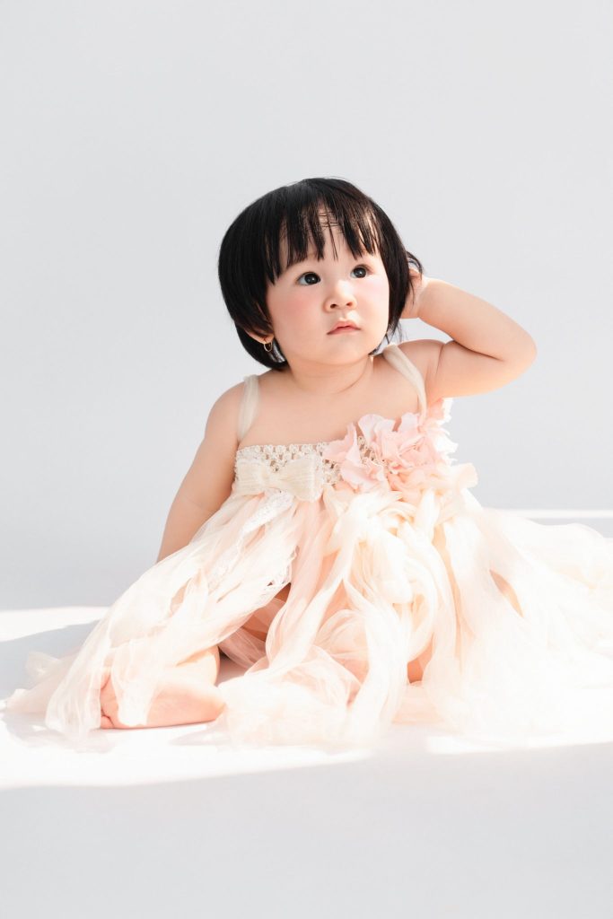 Bé cài hoa trắng trên tóc, đưa tay gần đầu và nhìn máy ảnh