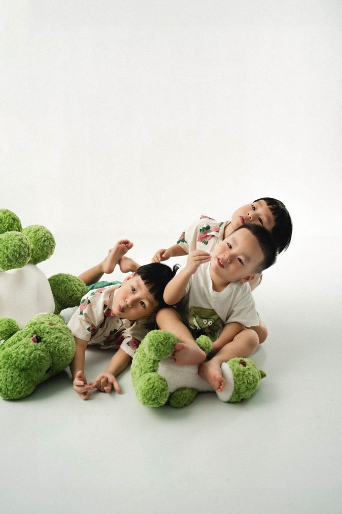 Ba bé mặc áo hoa ngồi cùng thú bông khủng long xanh