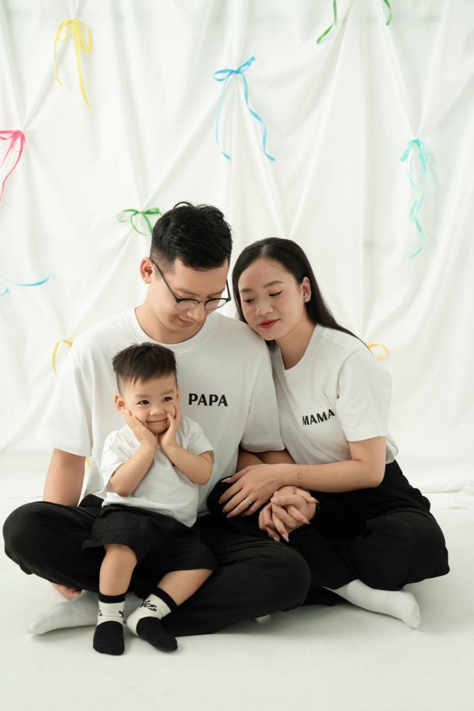 Ba mẹ và bé mặc áo trắng ngồi cùng nhau trước rèm màu pastel