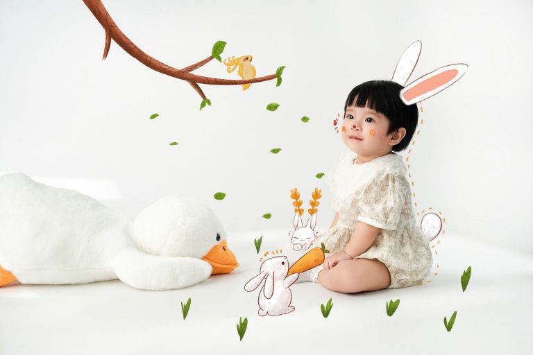 Bé mặc đồ kem ngồi cạnh ngỗng bông và hình tai thỏ