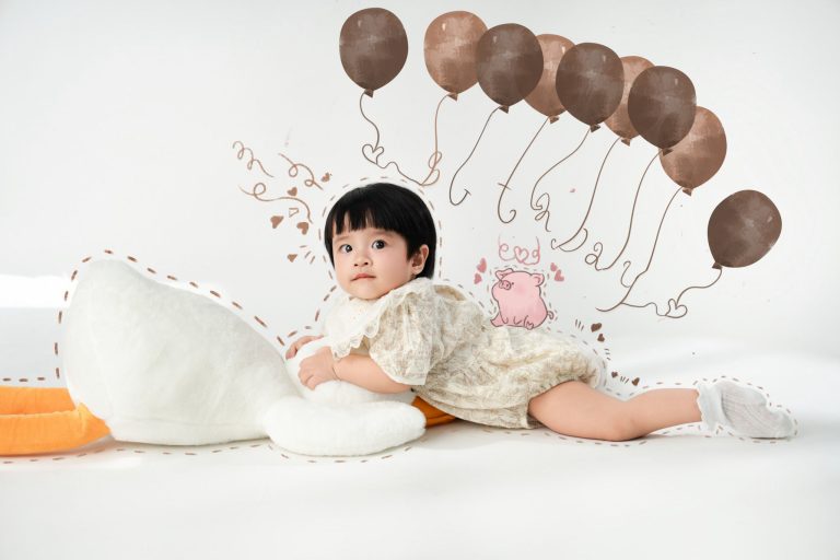 Bé nằm sấp cạnh ngỗng bông trắng và hình trang trí màu hồng