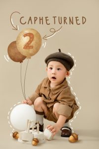 Bé mặc đồ nâu bên bóng và bảng sinh nhật hai tuổi