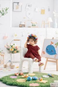 Bé mặc đồ đỏ sẫm đội vòng hoa, ngồi trên bục trắng bên tranh