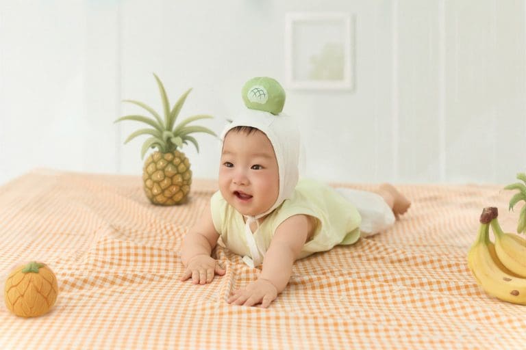 Bé nằm sấp trên nền sáng cùng các đạo cụ trái cây