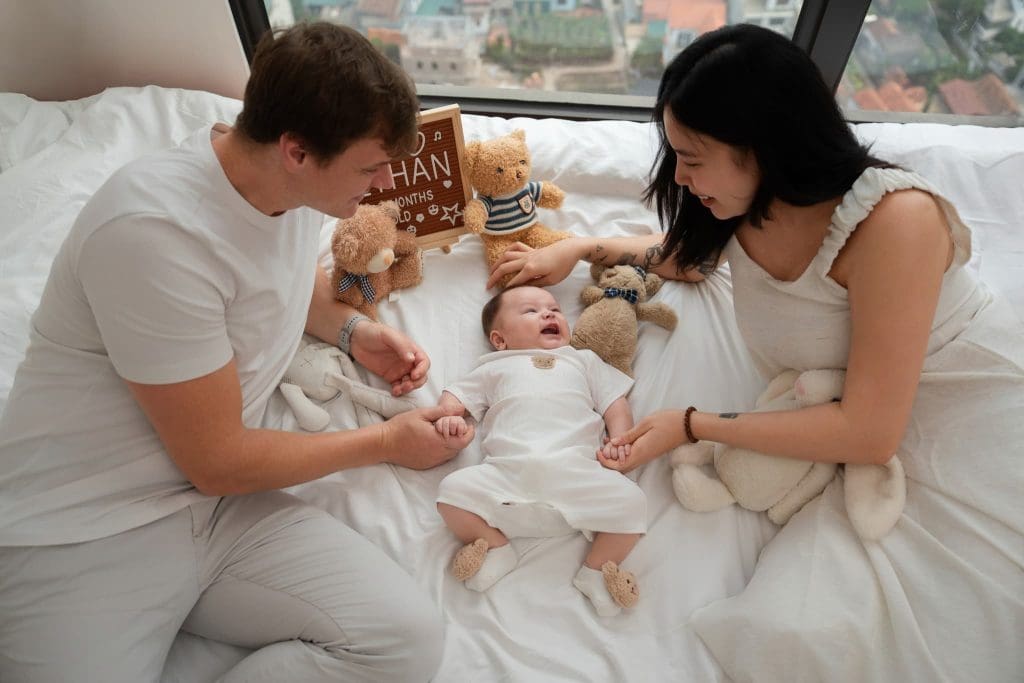 Ba mẹ ngồi trên giường bế bé mặc đồ trắng