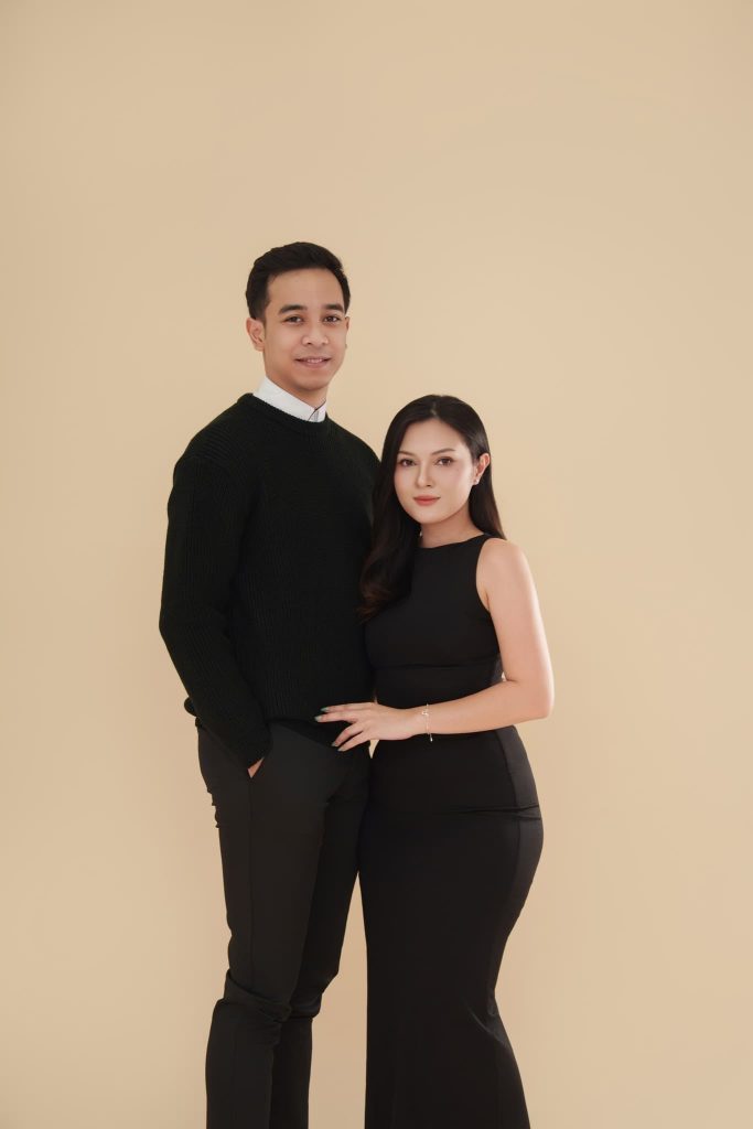 Bố đứng cạnh mẹ ngồi trên ghế, cả hai mặc đồ đen