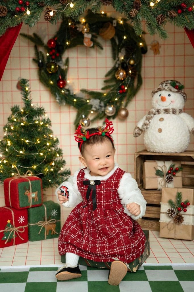 Bé mặc váy yếm kẻ đỏ đứng trên sàn ô xanh của bối cảnh Noel