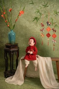 Em bé mặc áo dài đỏ đang khóc, ngồi trên ghế trước phông xanh.
