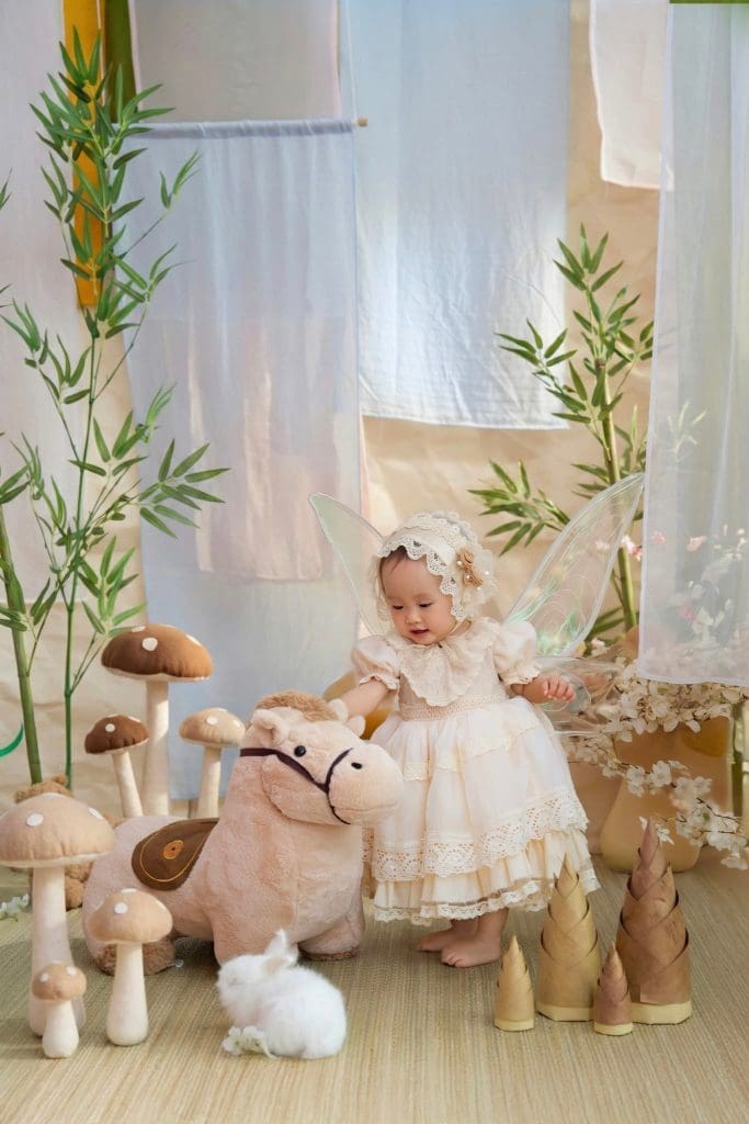 Bé đứng đặt tay lên ngựa bông cạnh màn vải trắng