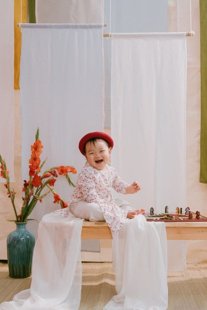 Bé mặc áo dài sáng màu ngồi trong bối cảnh hoa và rèm
