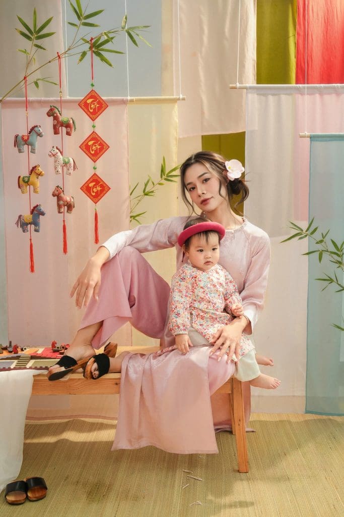 Mẹ bế bé mặc áo dài hồng trong bối cảnh hoa và rèm màu