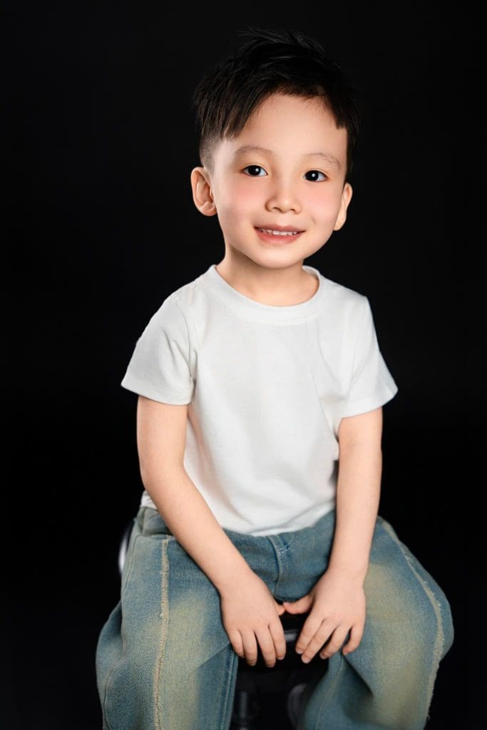 Bé ngồi cười trên nền đen, hai tay đặt phía trước