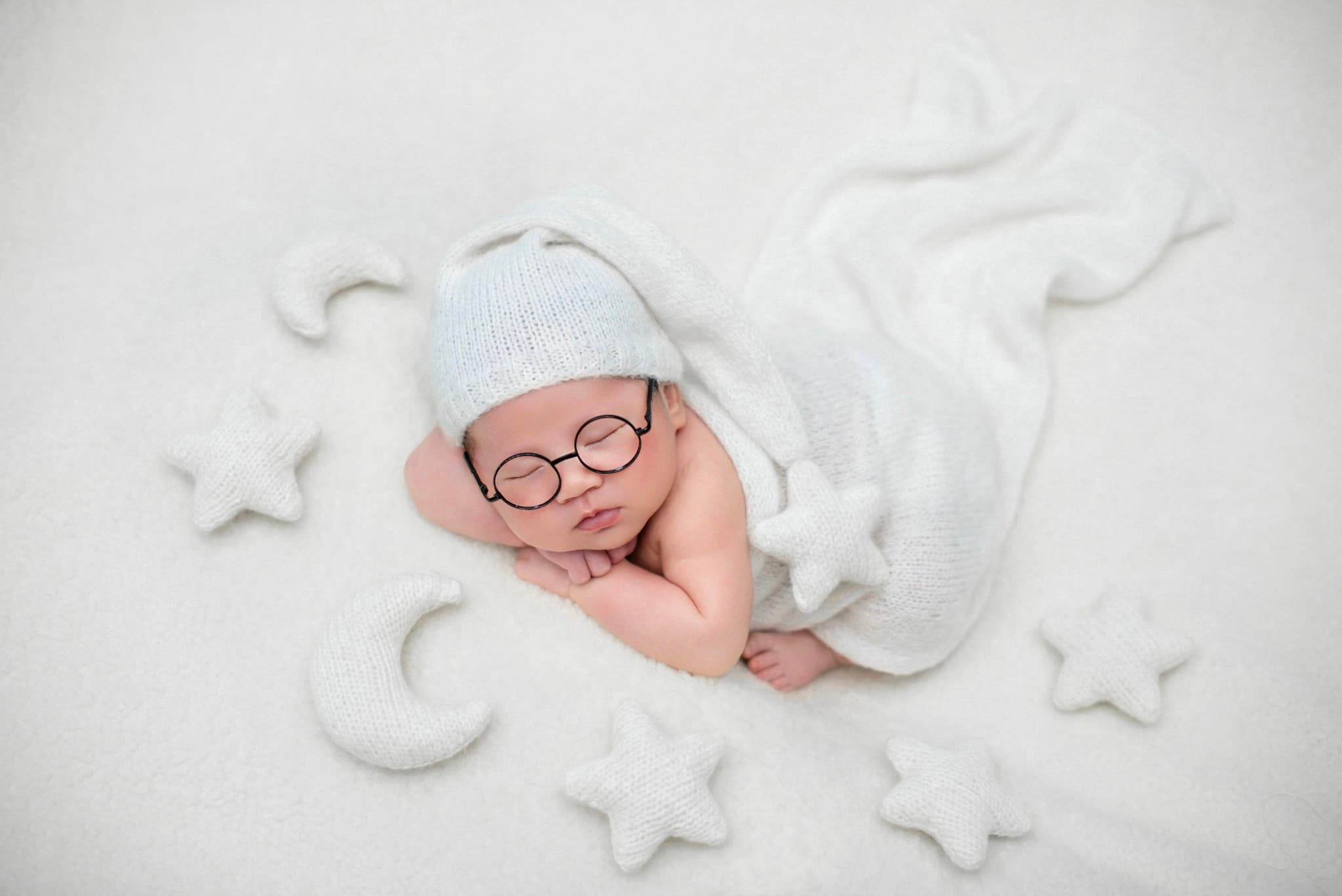 Bé sơ sinh đội mũ và đeo kính tròn ngủ trên nền trắng