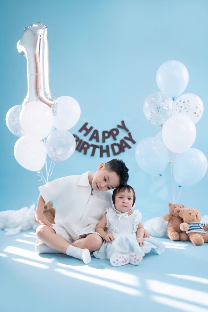 Bé mặc váy trắng ngồi trong bối cảnh sinh nhật xanh trắng